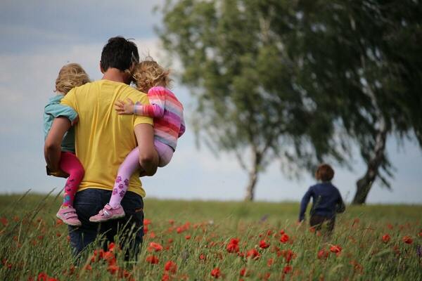 Father Holding Two Daughters In Field With Flowers