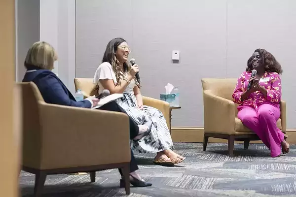 Three women seated in armchairs during a panel discussion. The woman in the center and the woman on the right are each holding a microphone and smiling as they speak. The moderator on the left is partially visible, taking notes. The setting appears to be an indoor event space.