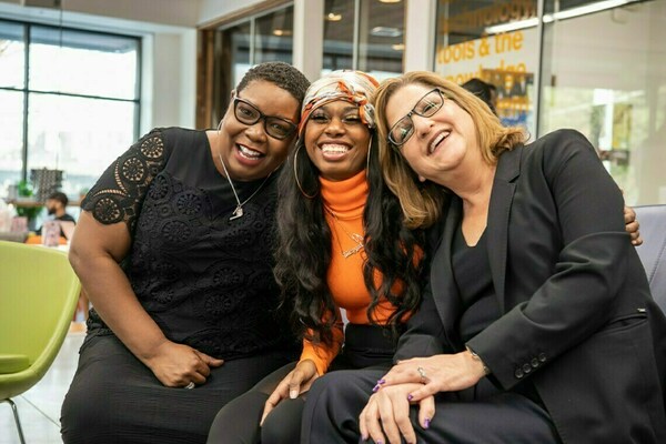 Three women are sitting closely together indoors, smiling warmly at the camera. The woman on the left wears glasses and a black lace top, the woman in the middle wears an orange turtleneck and a patterned headscarf, and the woman on the right wears glasses and a black blazer. They appear happy and relaxed, leaning in toward each other in a friendly, joyful moment.