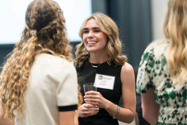 A woman wearing a black dress and a name tag that reads “Lexi Wetzel” smiles while holding a glass and talking with two other women at an event. The scene appears friendly and conversational, with all three engaged in discussion.