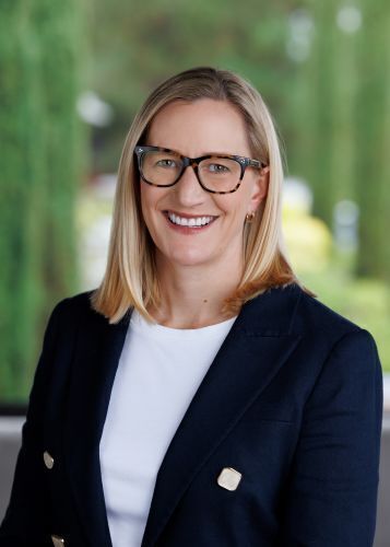 Smiling woman with medium length blonde hair wearing a navy blazer with gold buttons, a white top, and tortoise-shell glasses, against a blurred green background.