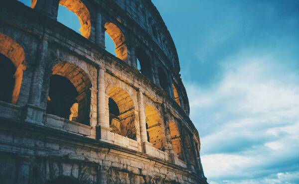 The Colosseum at dusk, with orange lights illuminating its arched facade against a deep blue sky.