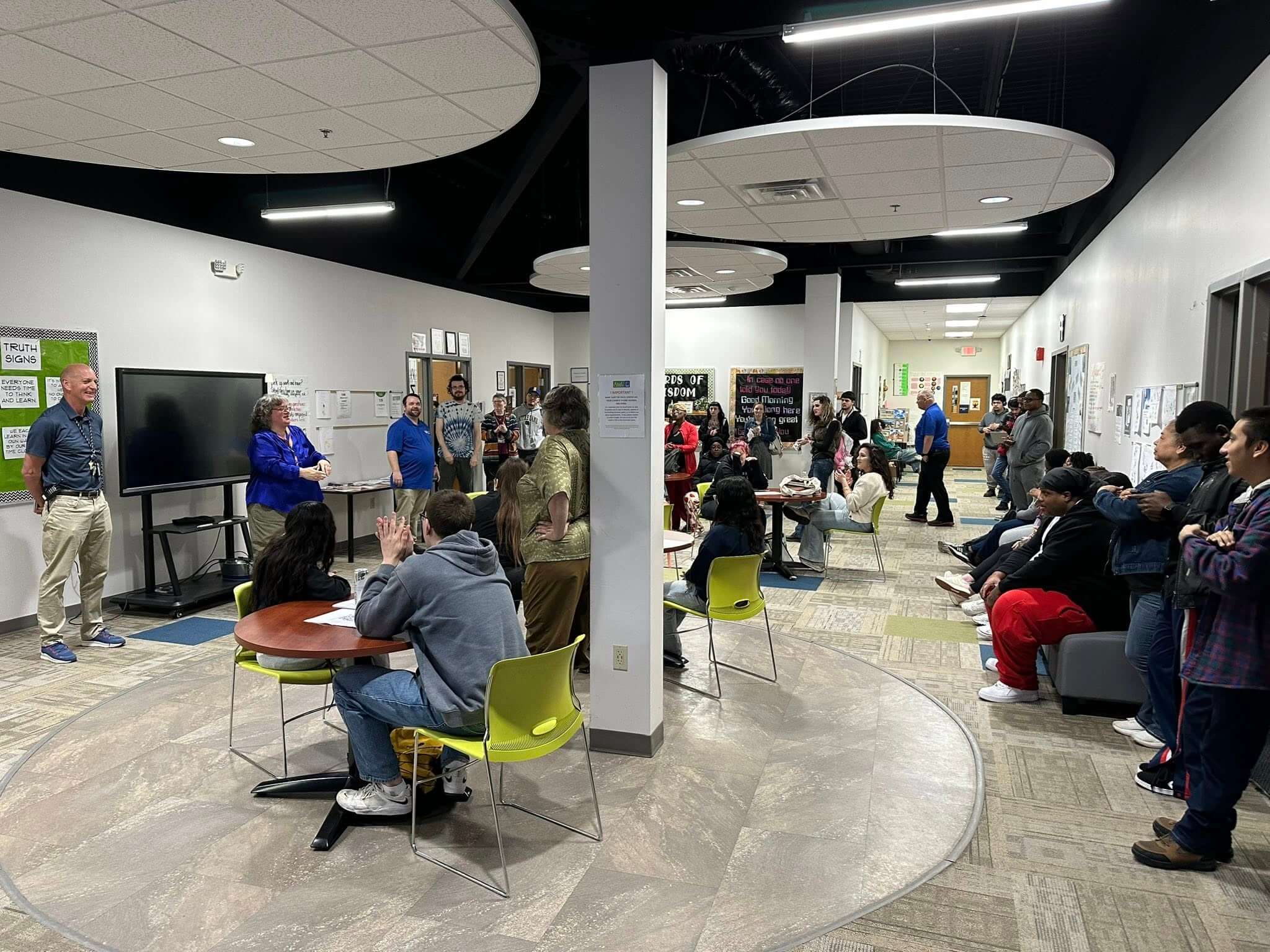 A man and woman present to a diverse group of adult students gathered in a bright, modern room. Students sit on lime chairs and stand.