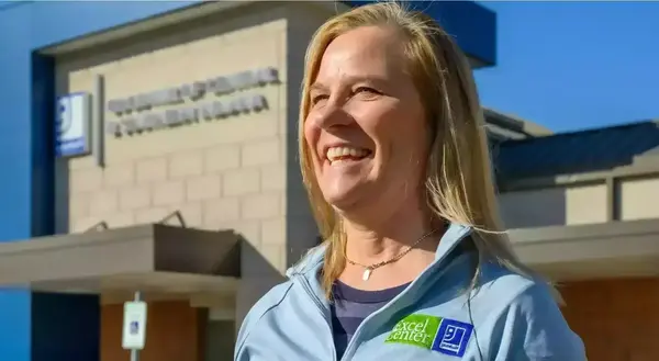 A blonde woman wearing a light blue Goodwill Excel Center jacket smiles broadly outside a Goodwill Excel Center building.