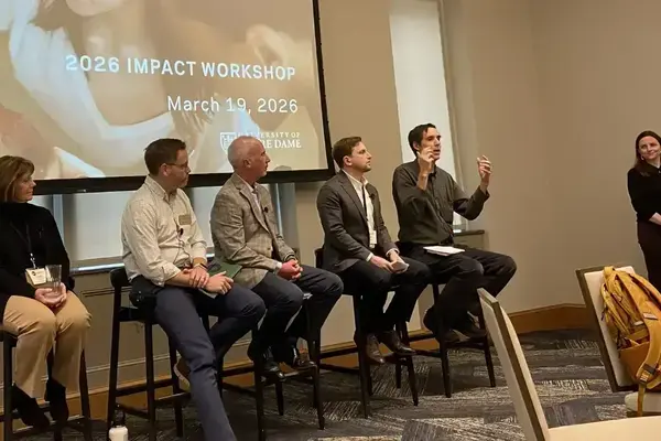 Five panelists sit on stools before a screen displaying '2026 IMPACT WORKSHOP March 19, 2026' and Notre Dame logo. One man gestures as he speaks. A woman stands by a podium moderating the panel.
