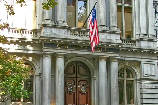 Gray stone Old City Hall building entrance with ornate brown wooden doors,  columns, and an American flag.