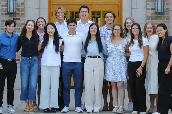 A group of smiling young men and women pose together outside a brick university building with a large wooden door.