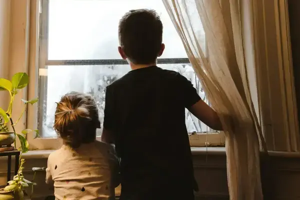 A young boy and girl look out a window at a snowy outdoor scene. The girl wears a light shirt with colorful drops. A house plant sits next to the window.