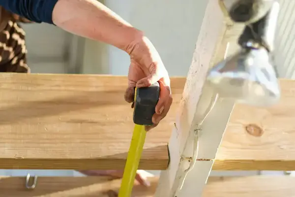 A person's hand, wearing a blue sleeve, measures a light wooden plank with a black and yellow tape measure.