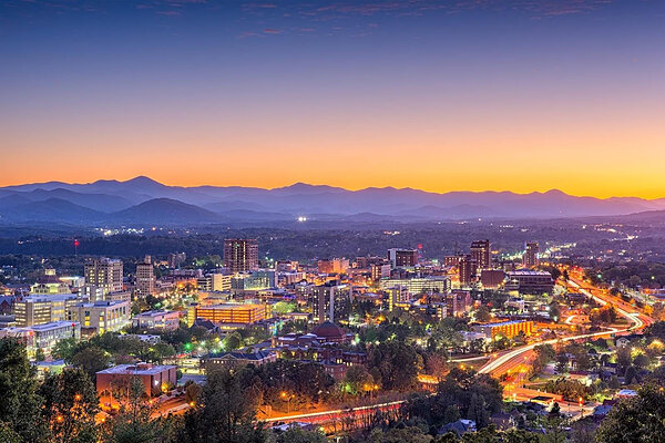 Asheville, NC cityscape at dusk with mountains, illuminated buildings, and light trails from vehicles.