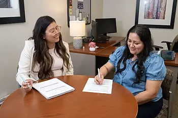 Two women at a round table, one in a white blazer, the other in a blue denim shirt, review documents and write.